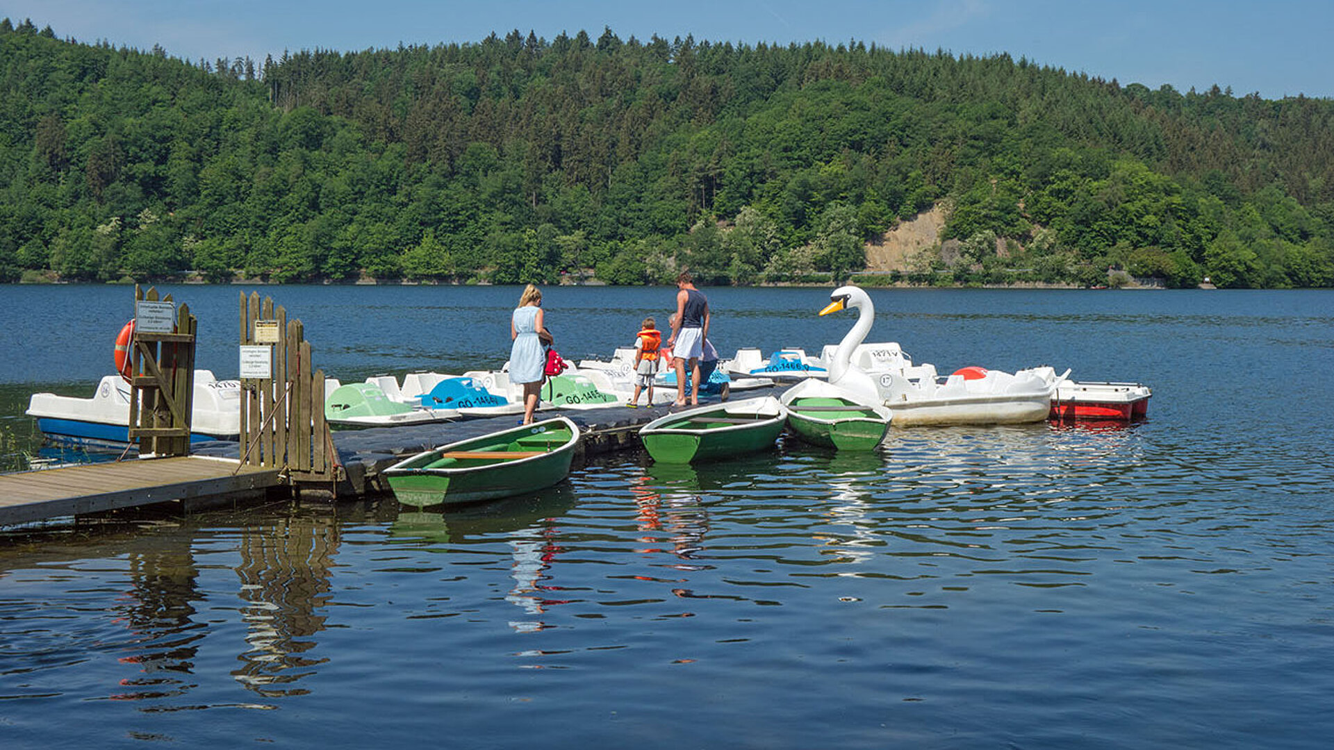 Familie am Bootsverleih mit Tretbooten und Ruderbooten auf einem ruhigen See.