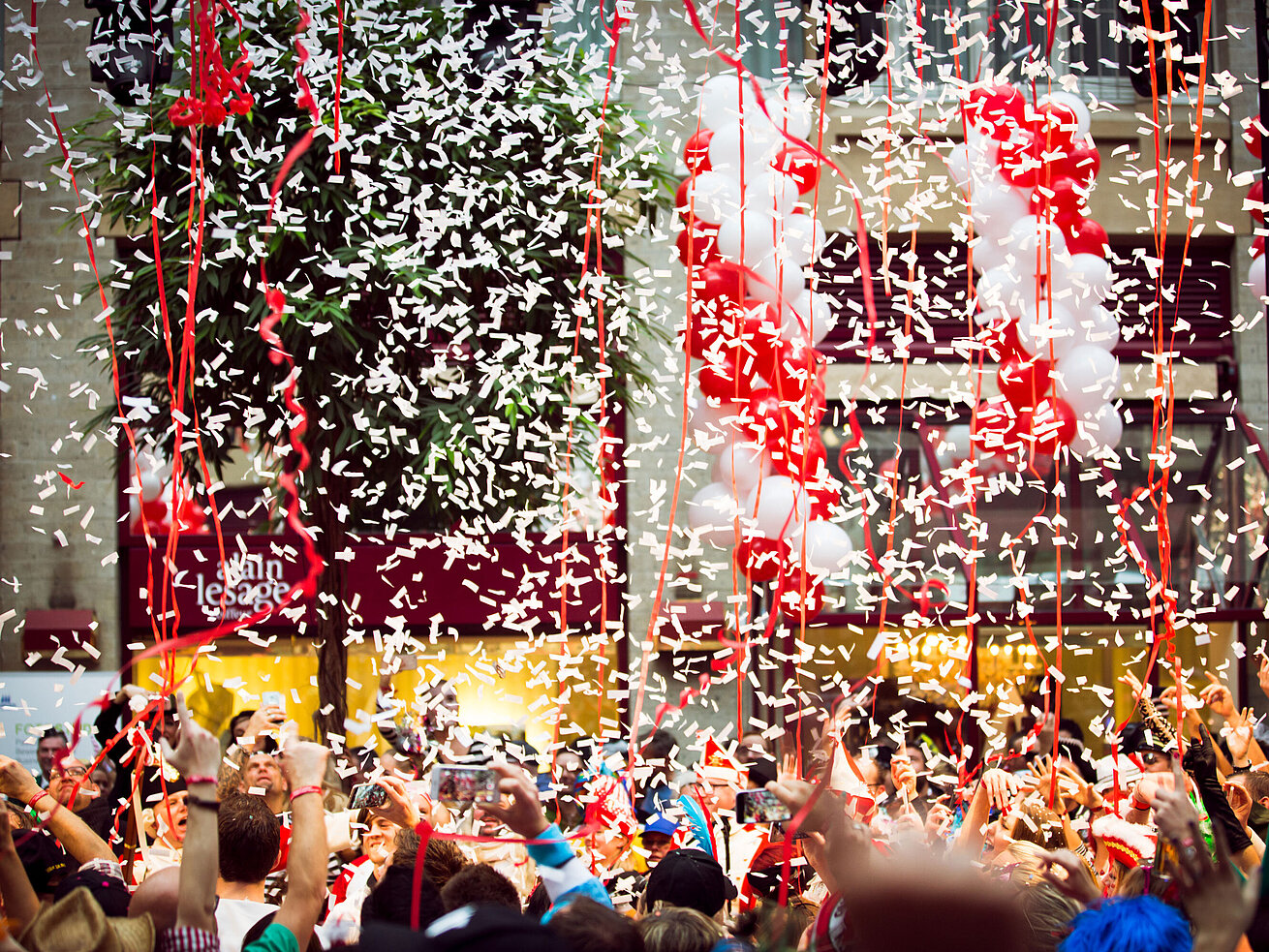 Konfetti und Luftschlangen regnen auf eine feiernde Menge herab, umgeben von rot-weißen Ballons bei einer Karnevalsparty.