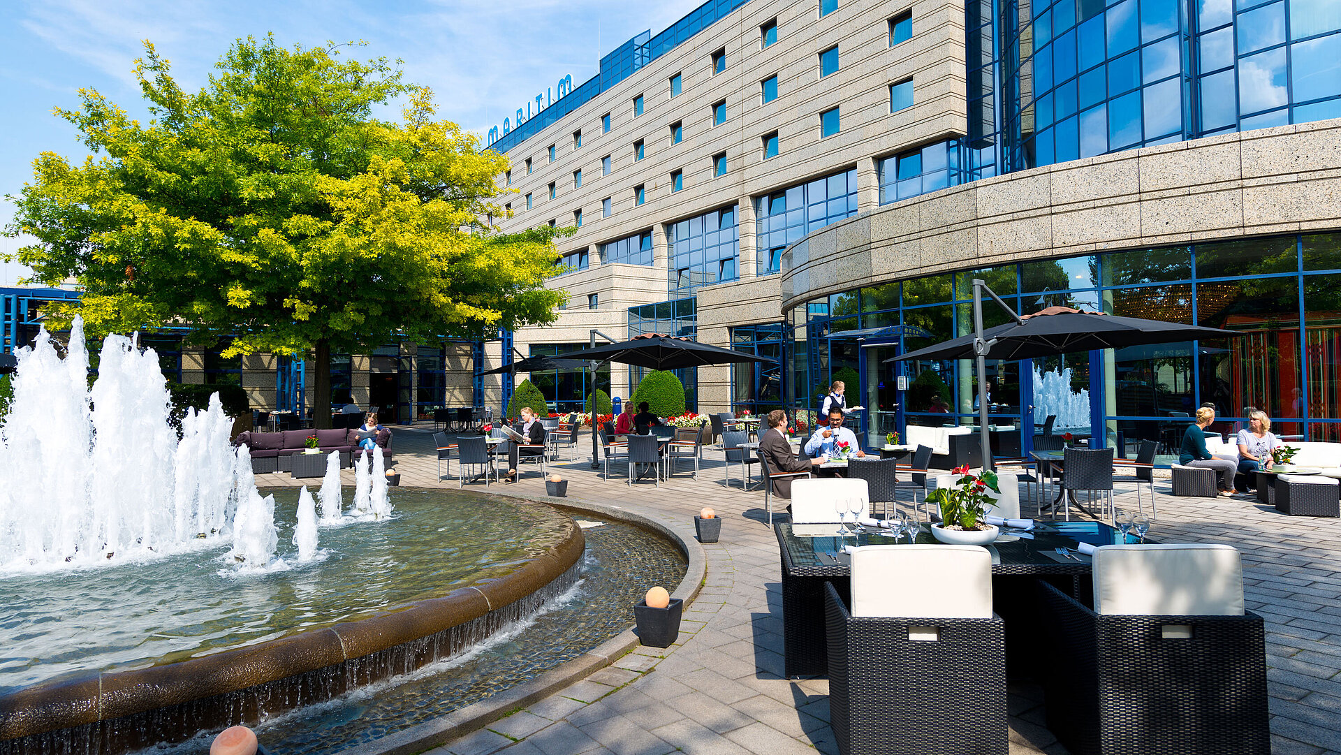 Terrasse Terrasse des Maritim Hotels Bonn mit Sitzbereichen, Springbrunnen und Gästen, die im Freien speisen und entspannen.