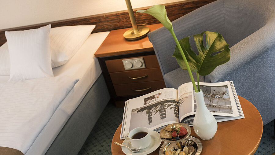 Details eines Zimmers im Maritim Hotel Kiel: Beistelltisch mit Vase, Buch und Snacks neben einem Bett und Sessel.