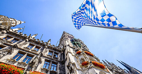 © fottoo - AdobeStock.com Bayerische Flagge über dem Marienplatz mit historischem Rathaus in München