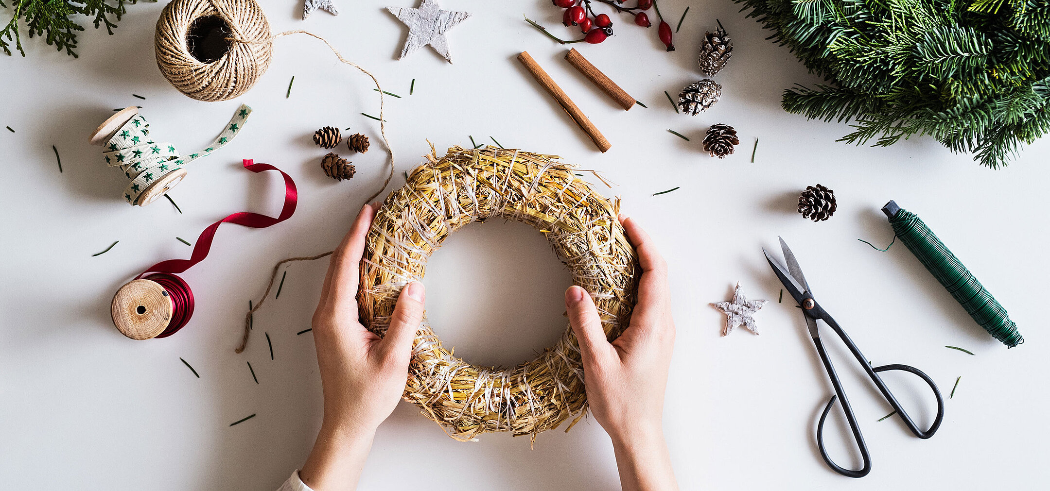 Adventskranz gestalten © Halfpoint - AdobeStock.com Hände gestalten einen Adventskranz auf einem Tisch mit Tannenzweigen, Schleifen und Bastelmaterial.