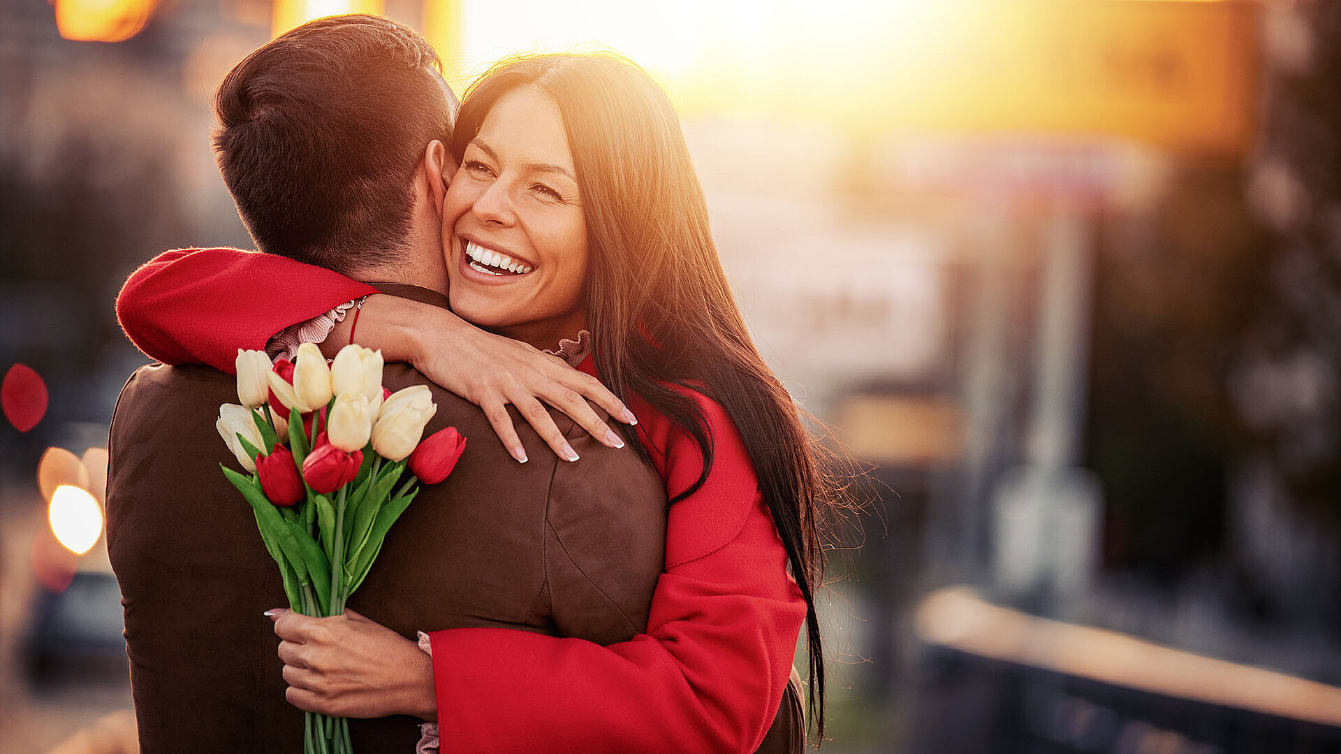 © Sanja 85 - AdobeStock.com Glückliches Paar umarmt sich bei Sonnenuntergang in der Stadt mit Blumenstrauß