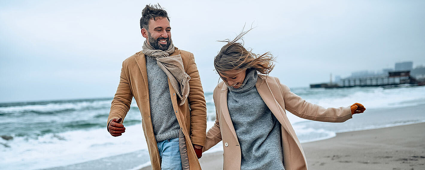 © Valerii Apetroaiei - AdobeStock.com Paar spaziert an einem windigen Tag in warmer Kleidung am Strand entlang.