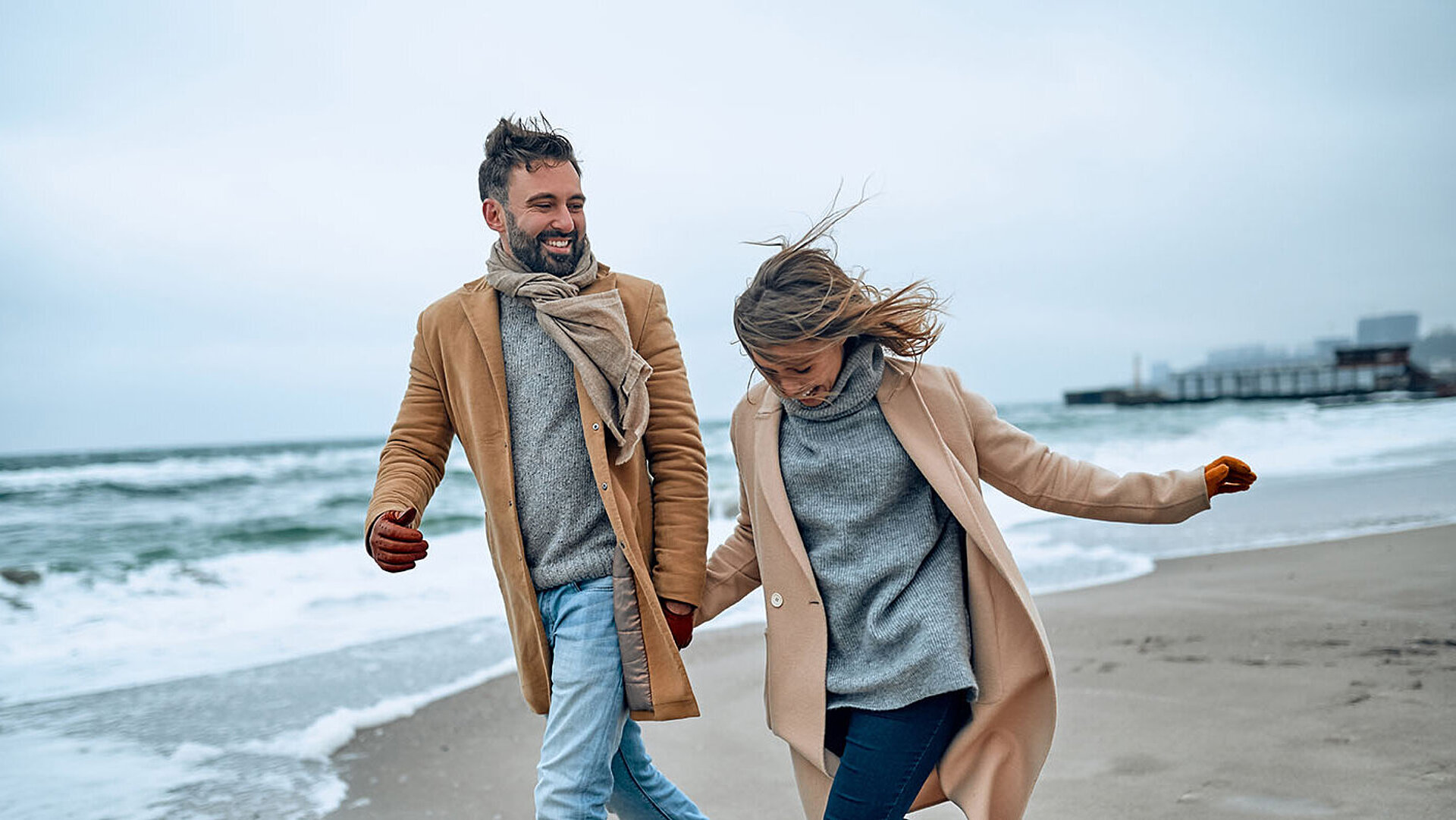 © Valerii Apetroaiei - AdobeStock.com Paar spaziert an einem windigen Tag in warmer Kleidung am Strand entlang.