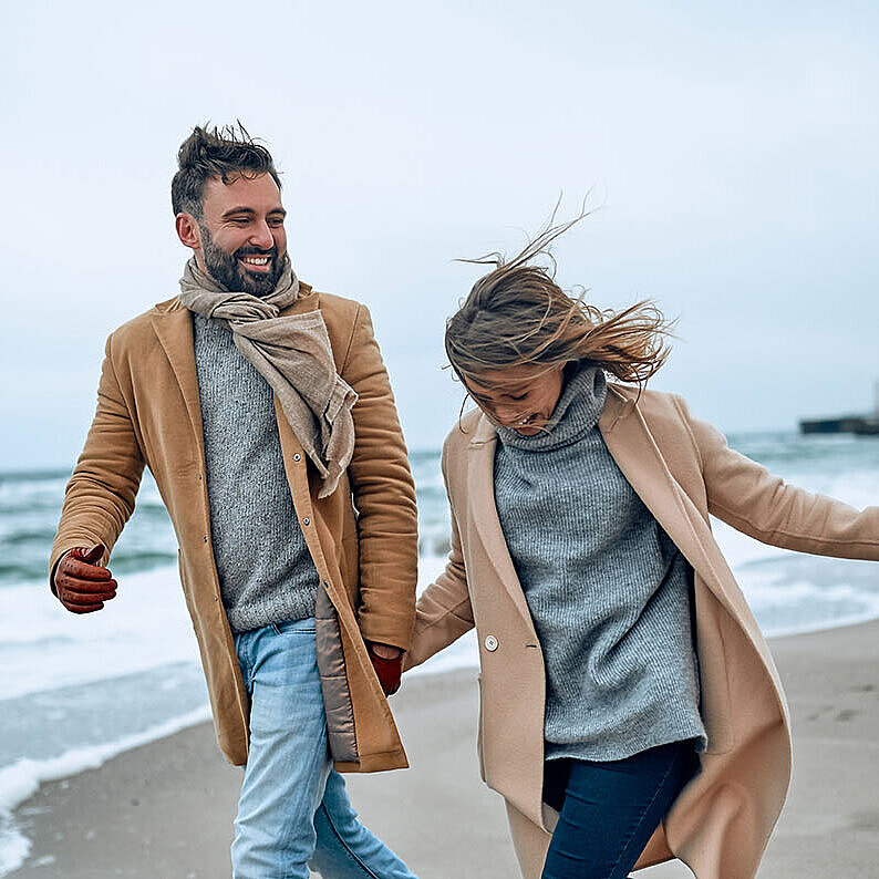 © Valerii Apetroaiei - AdobeStock.com Paar spaziert an einem windigen Tag in warmer Kleidung am Strand entlang.