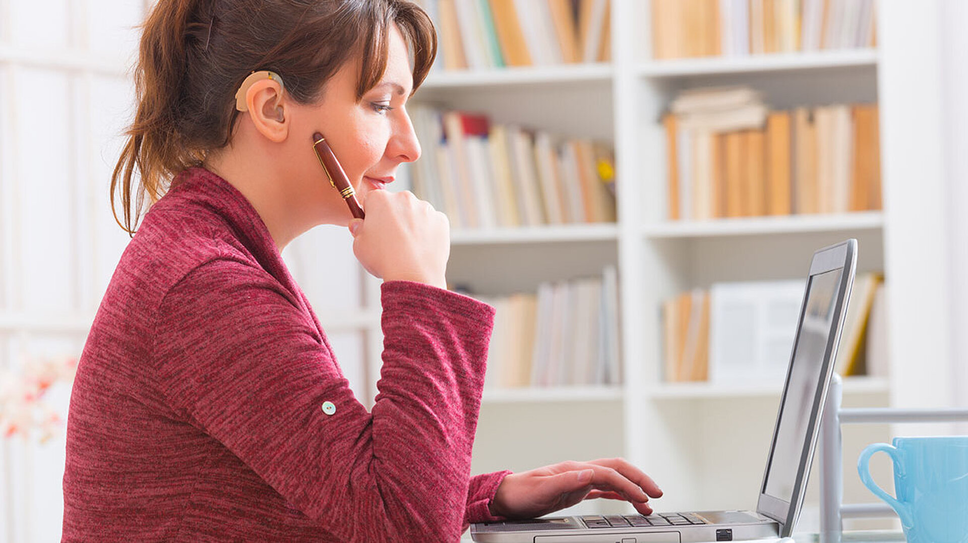 Frau mit Hörgerät arbeitet konzentriert am Laptop im Homeoffice vor einem Bücherregal.