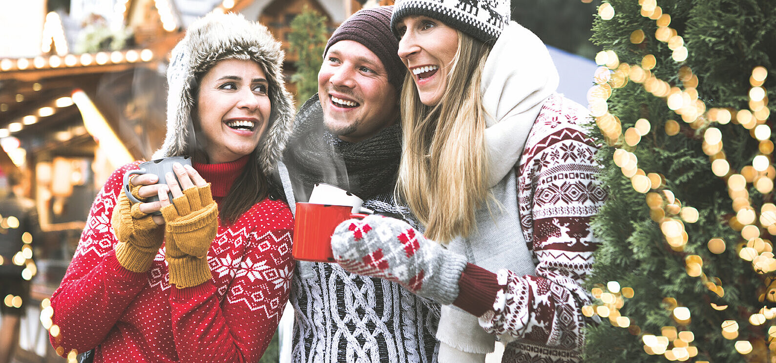 © drubig-photo - AdobeStock.com Drei lachende Freunde in Winterkleidung mit heißen Getränken auf einem Weihnachtsmarkt