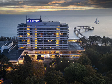 Abendaufnahme des Maritim Seehotels mit beleuchteter Seebrücke und einem Segelboot im Hintergrund bei Sonnenuntergang.