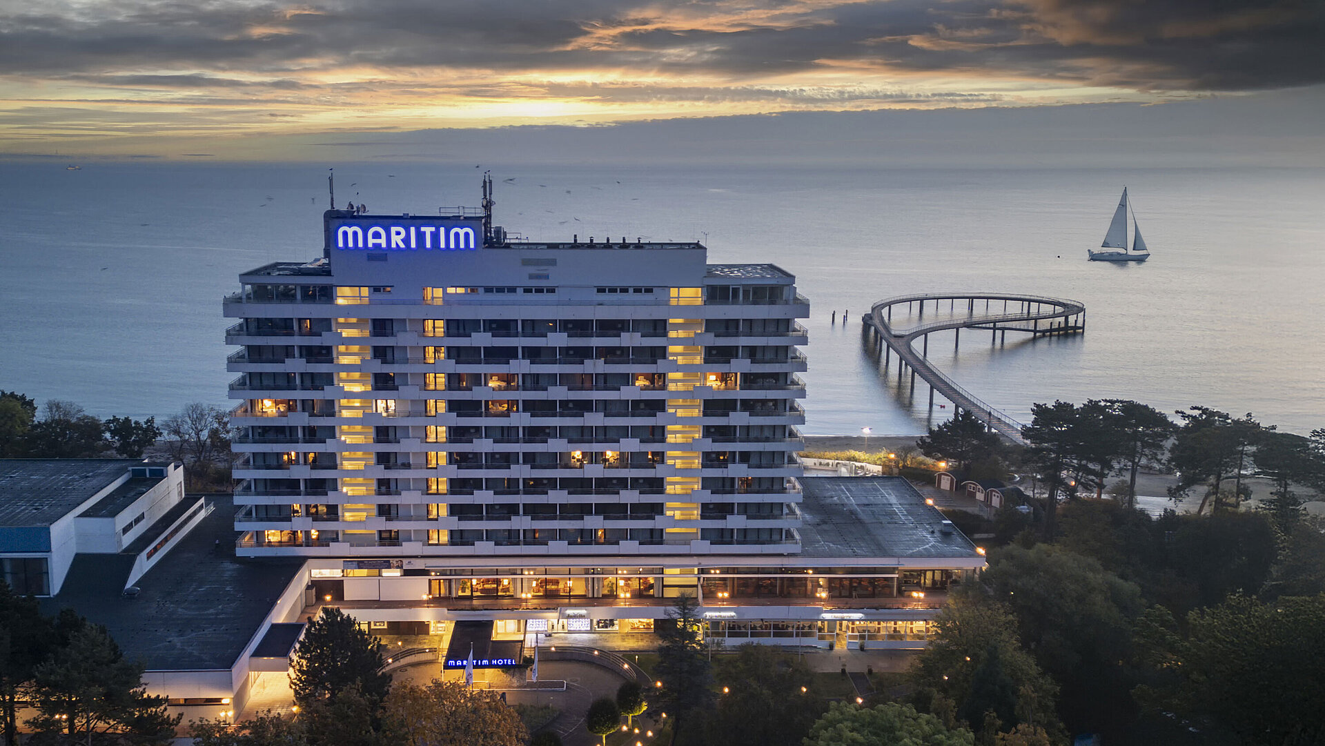 Außenansicht Abendaufnahme des Maritim Seehotels mit beleuchteter Seebrücke und einem Segelboot im Hintergrund bei Sonnenuntergang.