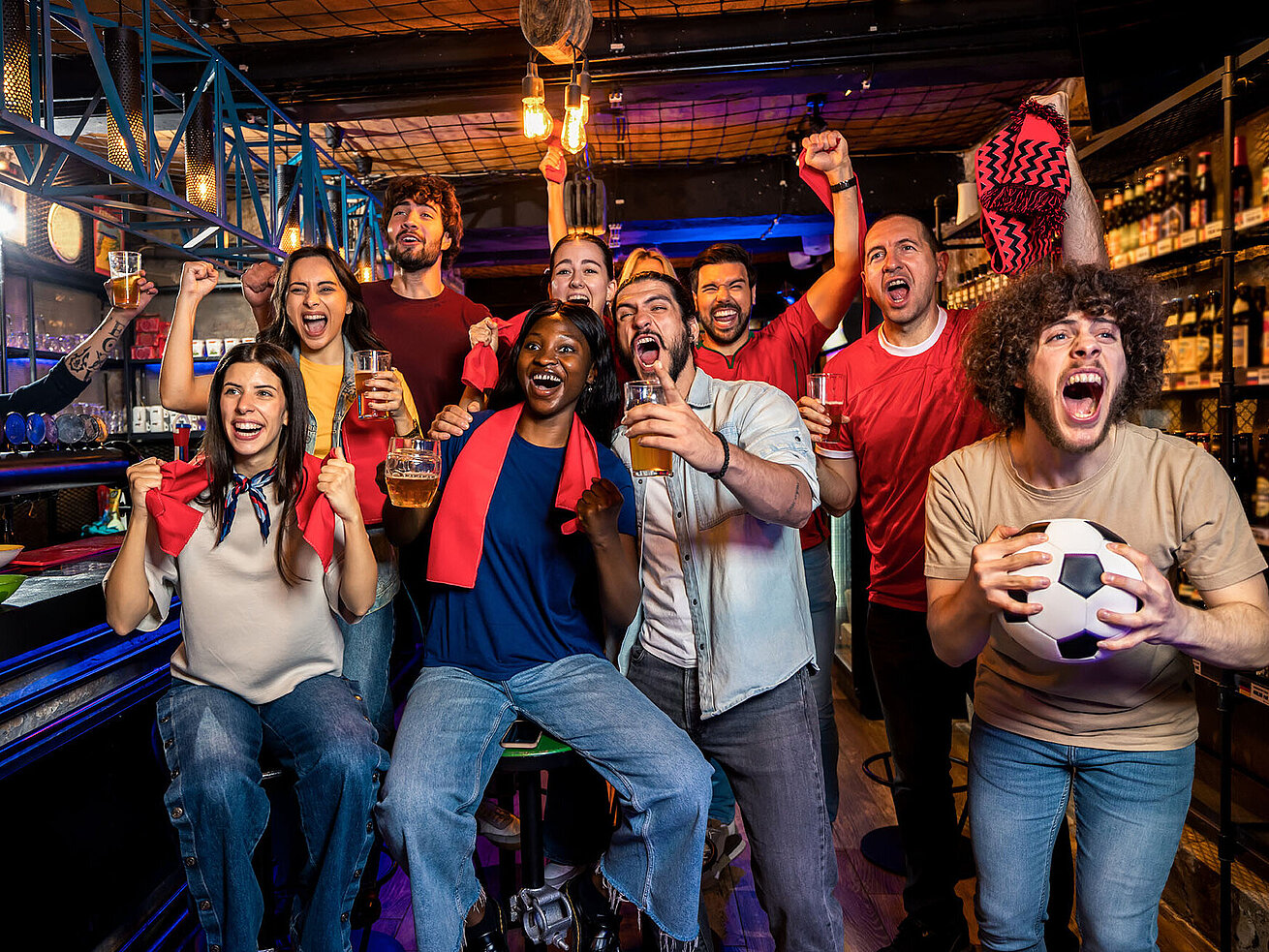 © Mediteraneo - AdobeStock_2017763771 Viele Leute jubeln zusammen in einer Bar. Sie tragen Fußball-Accessoires