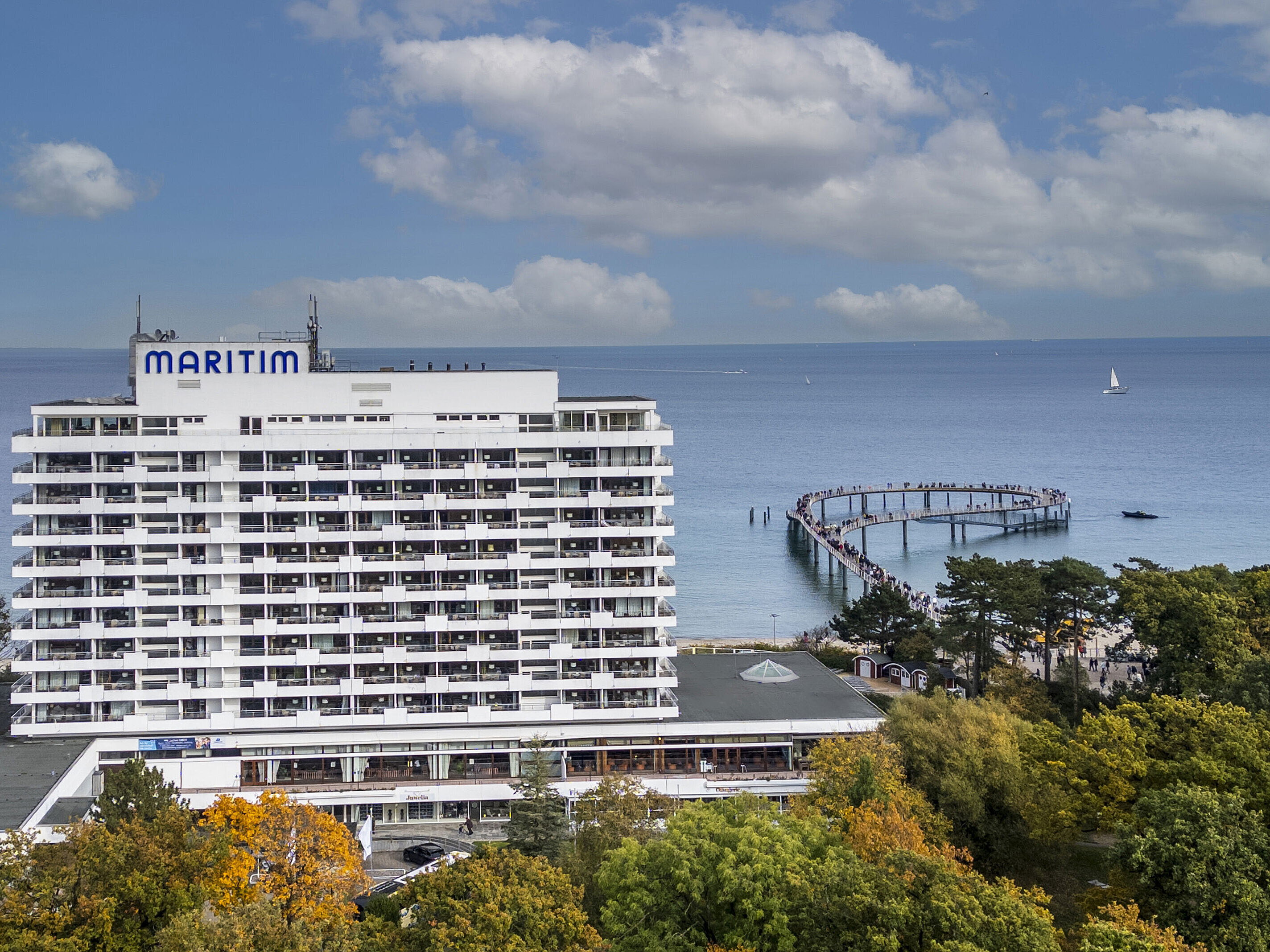 Maritim Seehotel Timmendorfer Strand mit Blick auf die Seebrücke und die Ostsee, umgeben von Bäumen und blauem Himmel.