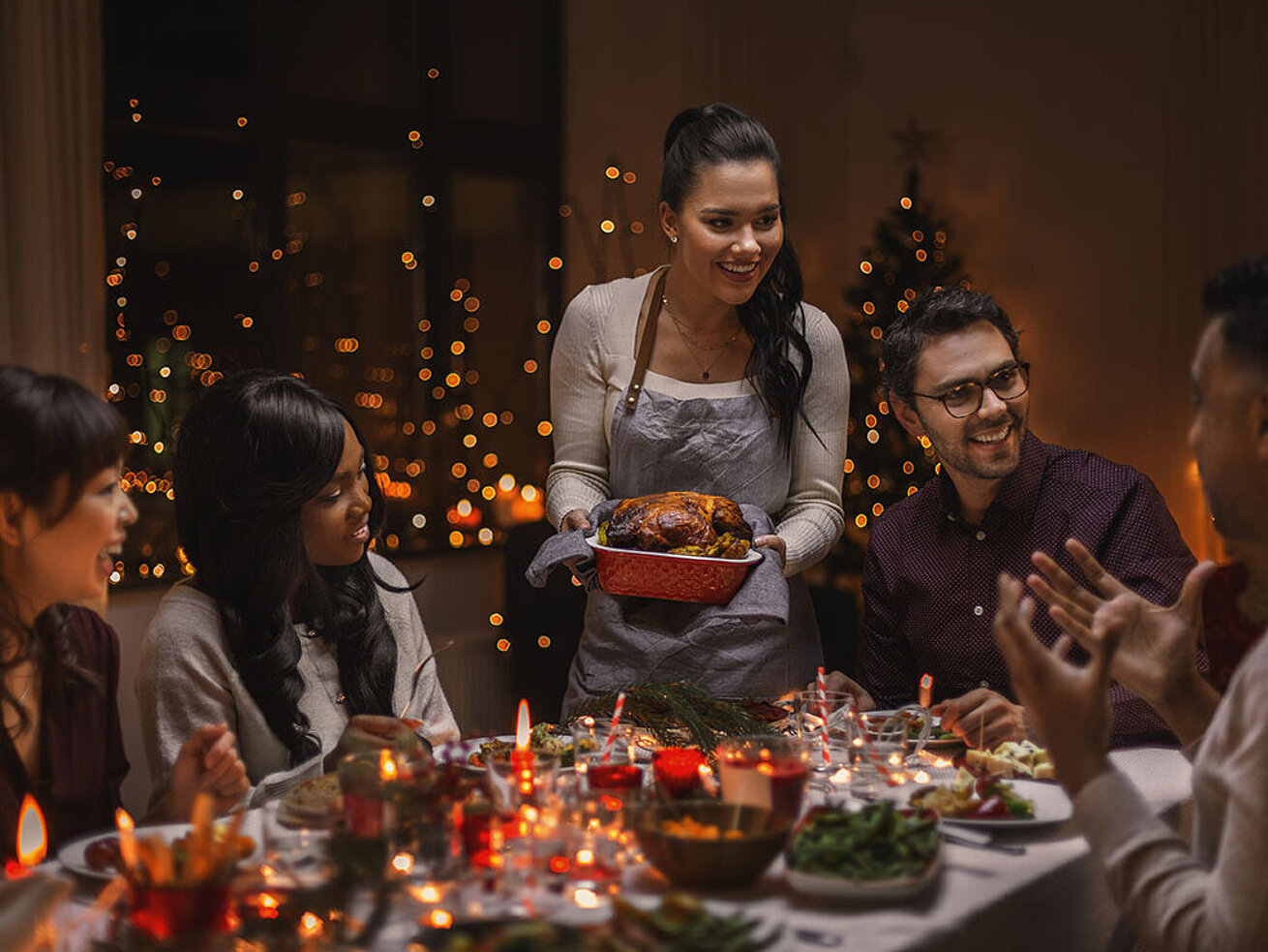 Festlich gedeckter Tisch mit Freunden beim Weihnachtsessen und einer Frau, die den Braten serviert.