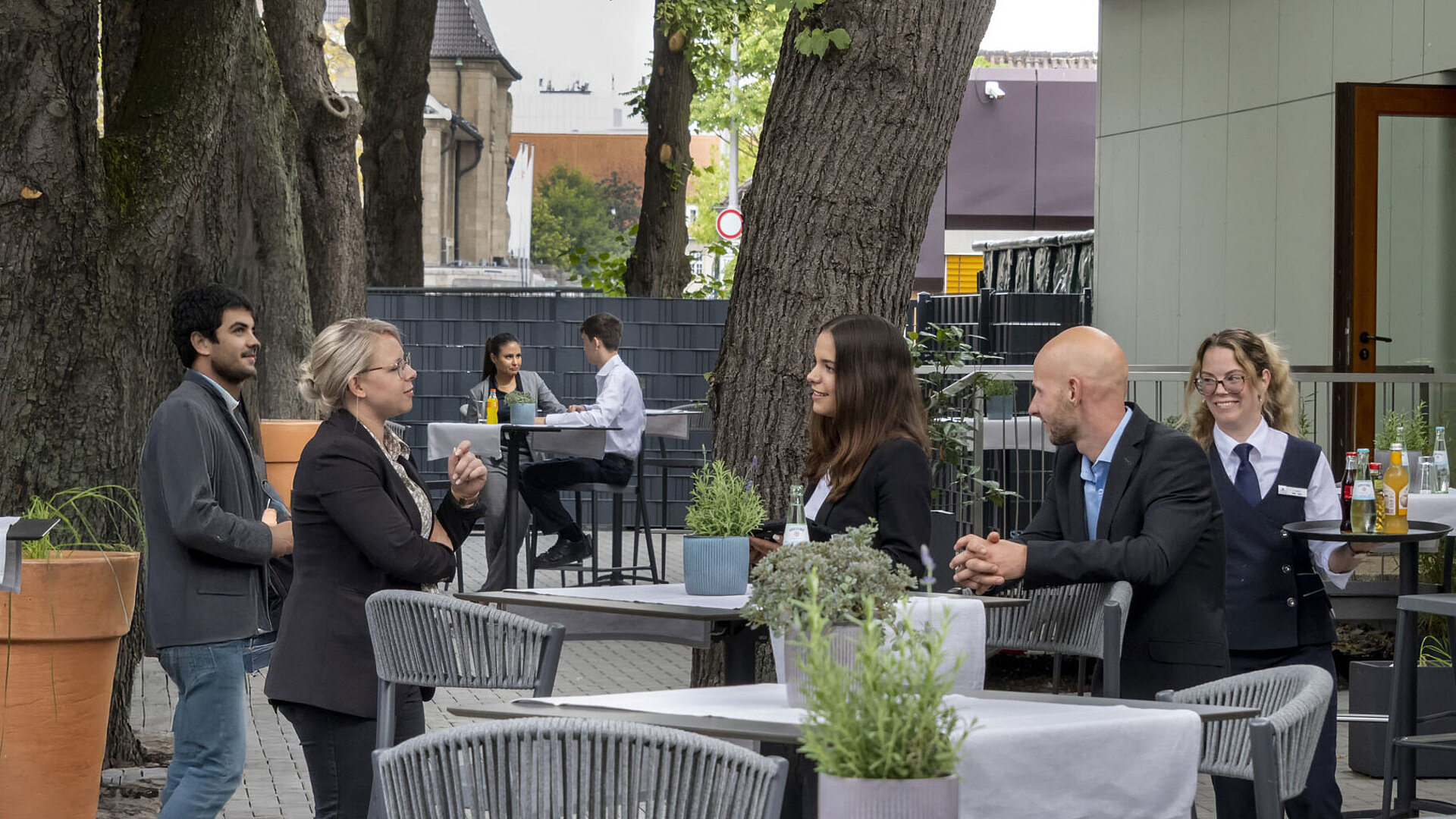 Moderne Terrasse im Maritim Hotel Darmstadt mit Gästen, die entspannen, Bäumen im Hintergrund und stilvollen grauen Möbeln.