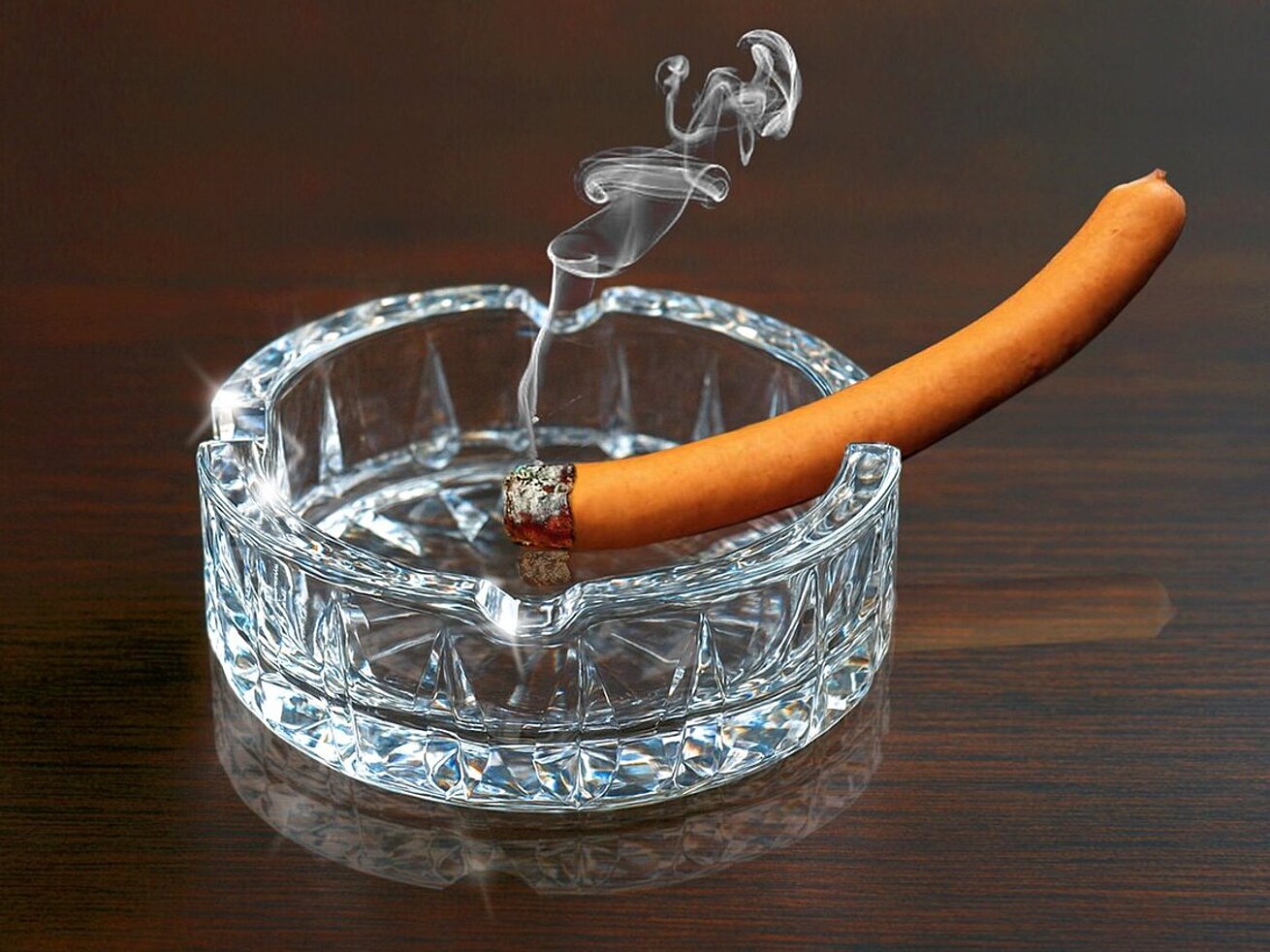 Wurst in einem Glasaschenbecher mit brennendem Ende, es raucht, humorvolle und surreale Bildkomposition auf Holztisch