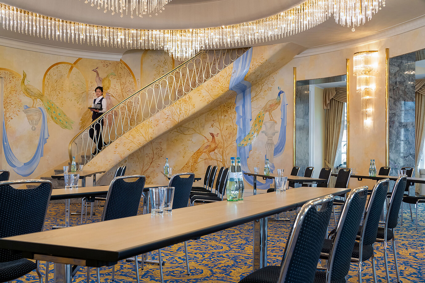 Fürst-von-Pückler-Saal mit Reihenbestuhlung, künstlerischer Wandgestaltung, großer Treppe und Servicekraft auf der Treppe.