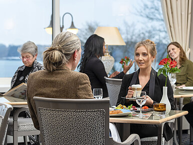Gäste im Restaurant Bellevue im Maritim Hotel Kiel, entspannte Gespräche bei Getränken und Speisen mit Blick auf das Meer.