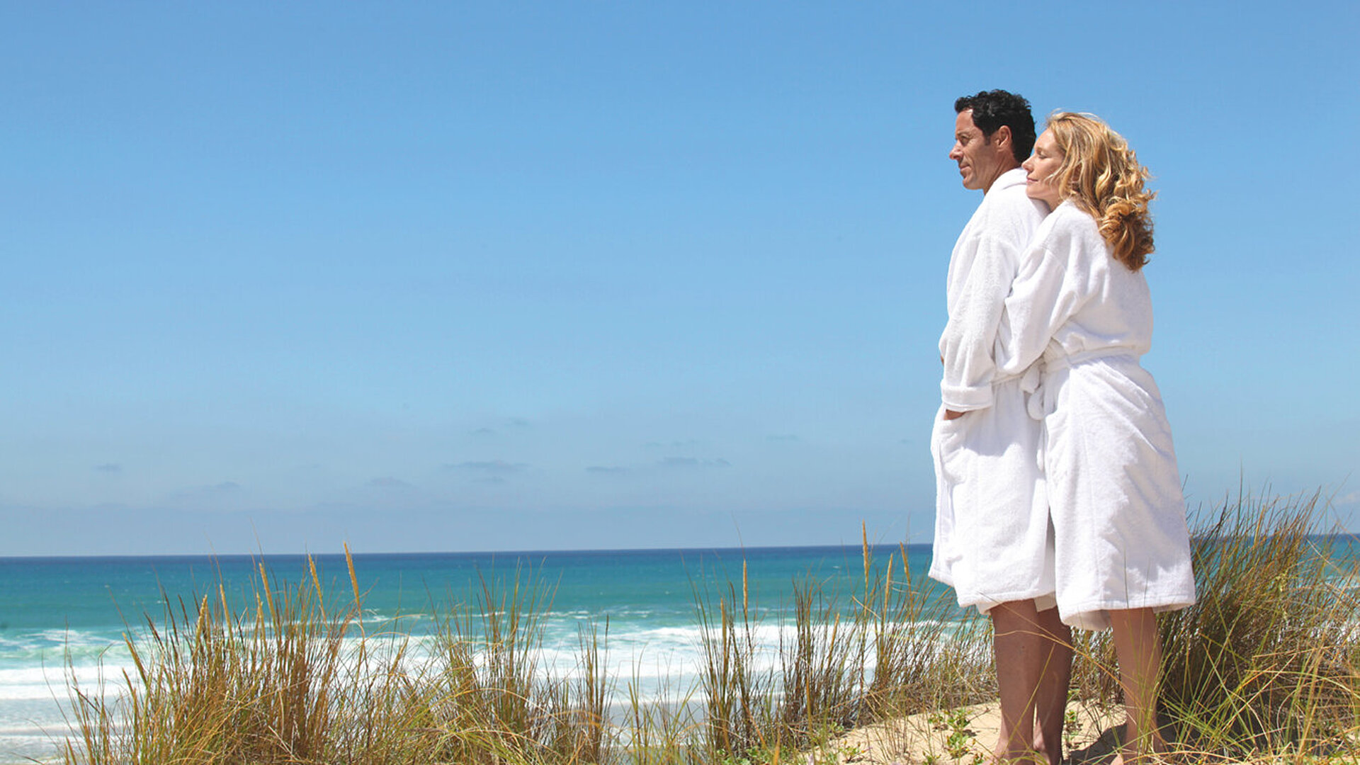 © auremar - AdobeStock.com Paar in weißen Bademänteln steht am Strand und blickt auf das Meer bei sonnigem Wetter.