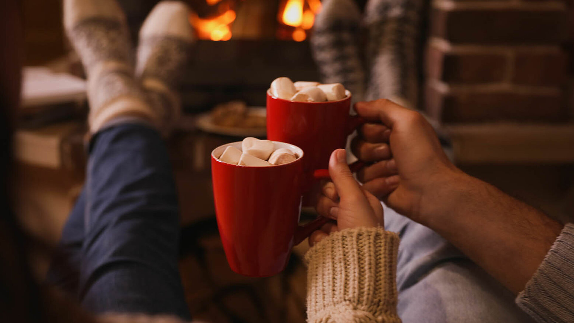 Zwei Personen entspannen am Kamin und halten rote Tassen mit Marshmallows in einer gemütlichen Winteratmosphäre.