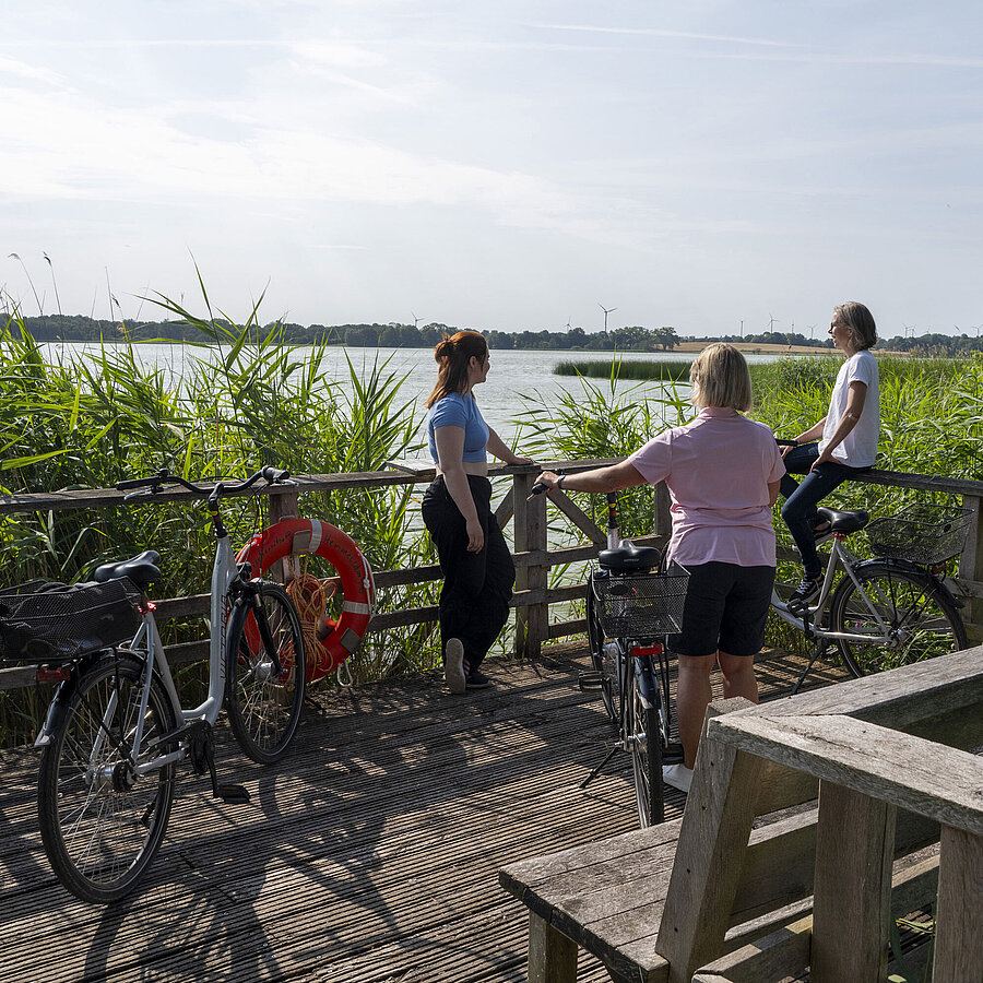 Drei Frauen machen eine Pause auf einer Fahrradtour an einem Steg mit Blick auf den See beim Maritim Timmendorfer Strand.