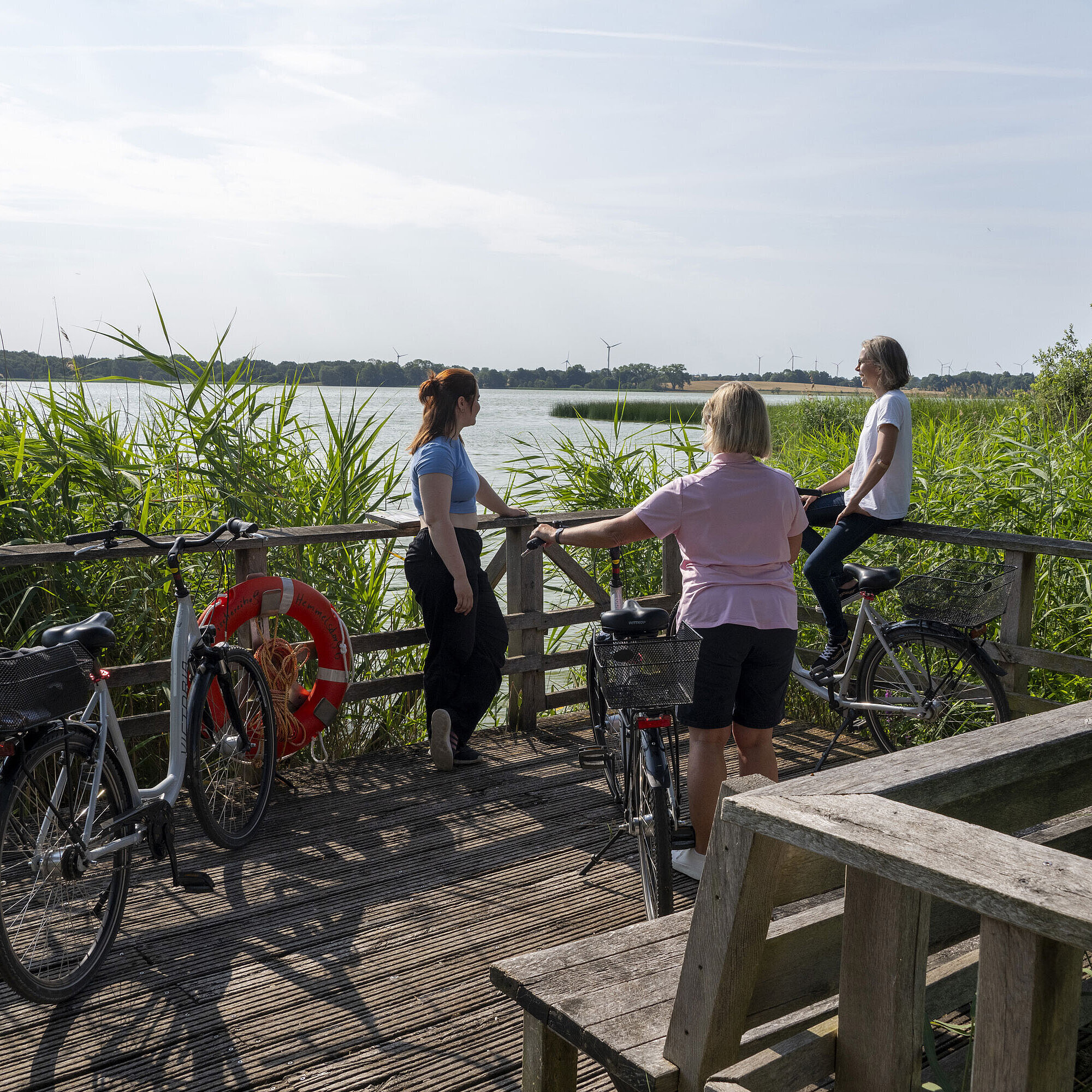 Drei Frauen machen eine Pause auf einer Fahrradtour an einem Steg mit Blick auf den See beim Maritim Timmendorfer Strand.