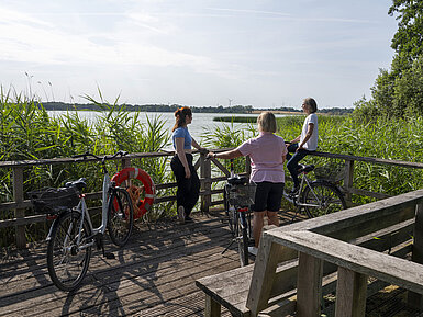 Drei Frauen machen eine Pause auf einer Fahrradtour an einem Steg mit Blick auf den See beim Maritim Timmendorfer Strand.