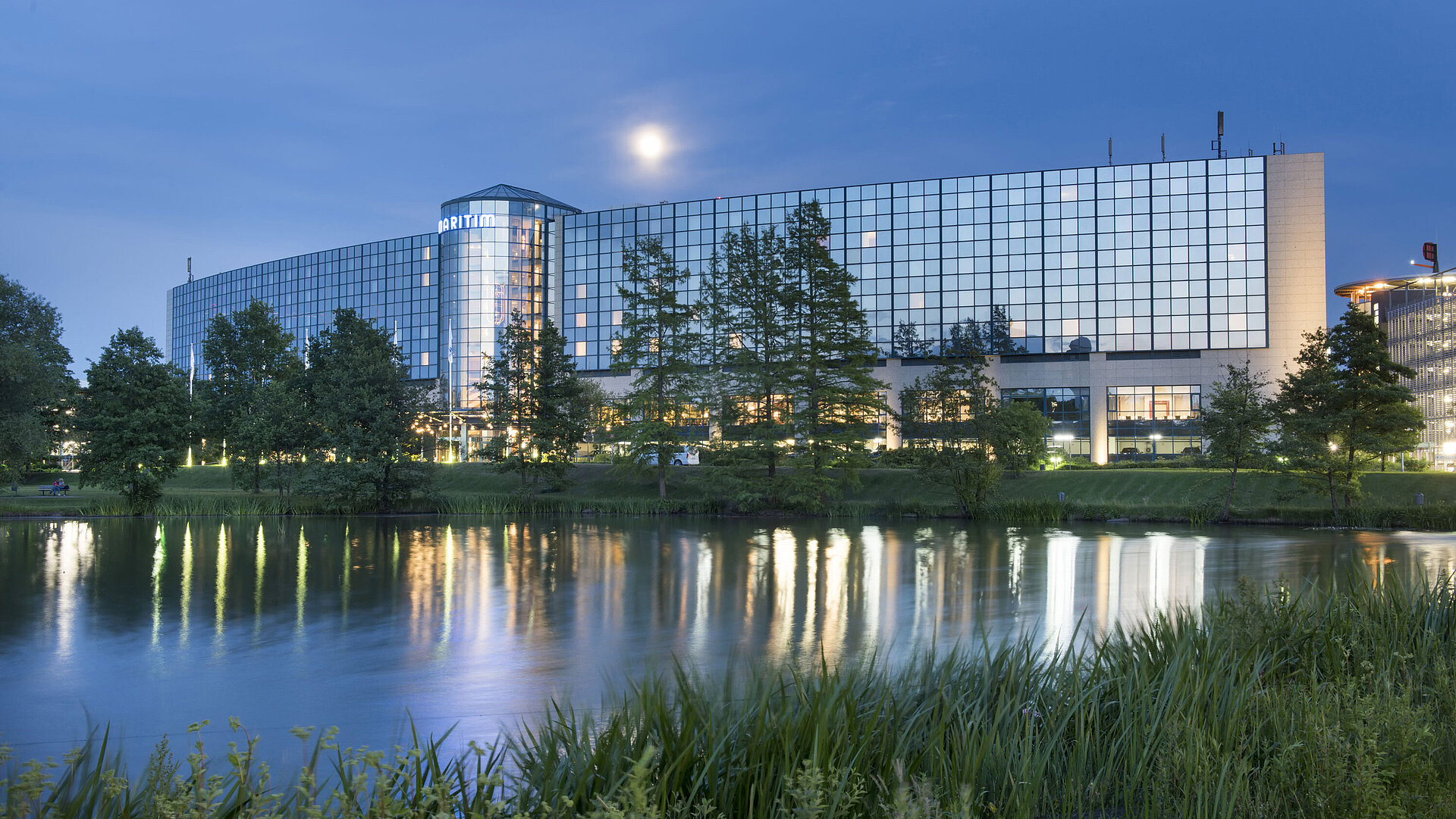 Beleuchtetes Maritim Hotel Hannover mit Glasfassade und Seespiegelung in der Abenddämmerung.