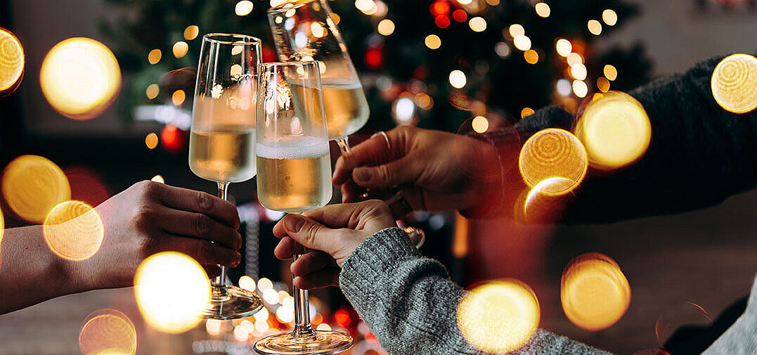 © Gregory Lee - AdobeStock.com Drei Personen stoßen mit Sektgläsern vor einem festlich beleuchteten Weihnachtsbaum an