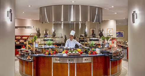 Küchenchef an der Show-Cooking-Station im Maritim proArte Hotel Berlin, umgeben von frischem Gemüse und Kräutern.