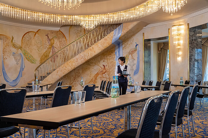 Fürst-von-Pückler-Saal mit Reihenbestuhlung, auffälliger Wandgestaltung, großer Treppe und Servicekraft im Raum.