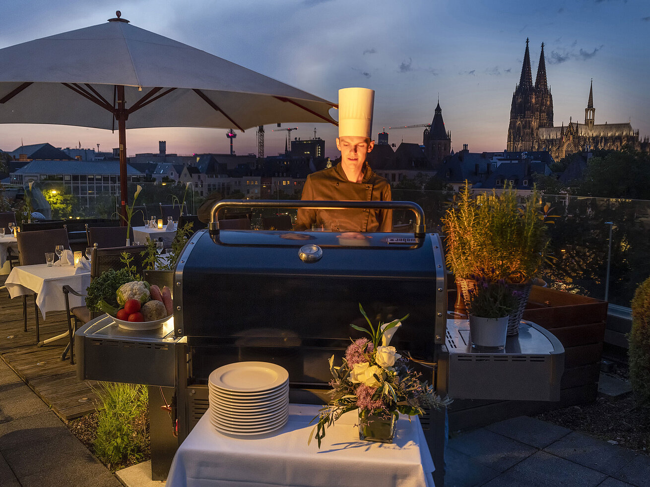 Ein Koch grillt abends auf der Dachterrasse des Maritim Hotels Köln mit Blick auf den beleuchteten Kölner Dom im Hintergrund.
