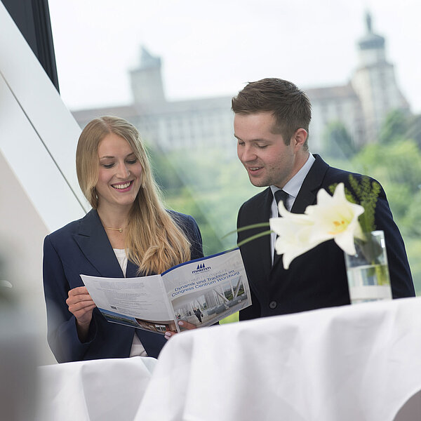 Zwei Personen im Gespräch im Maritim Hotel Würzburg mit Broschüre vor großem Fenster mit Festungsblick