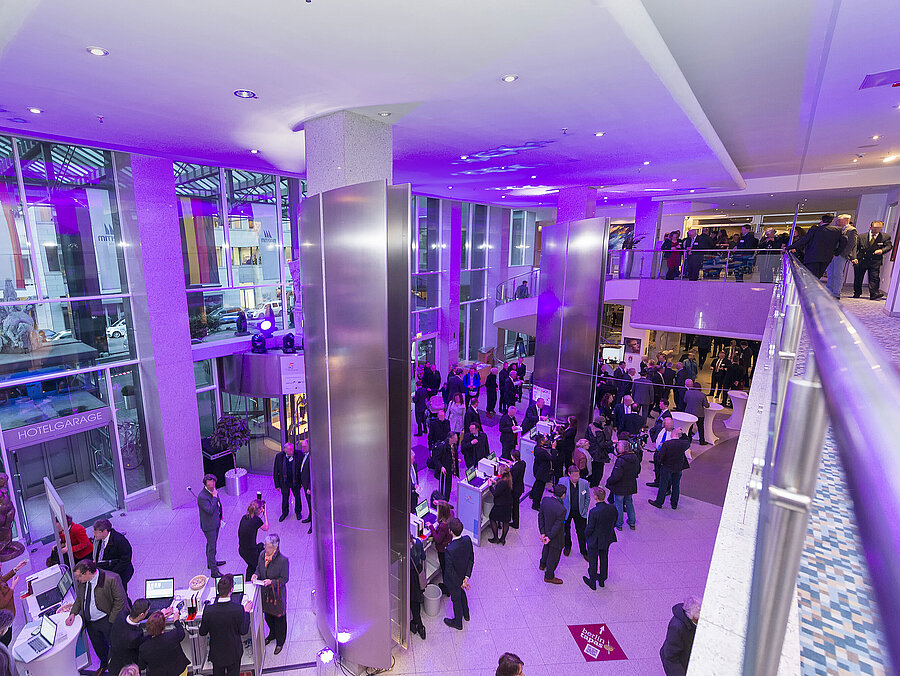 Eine Abendveranstaltung im Maritim Hotel Berlin. Die Lobby ist in lila Licht getaucht, und viele Gäste unterhalten sich.