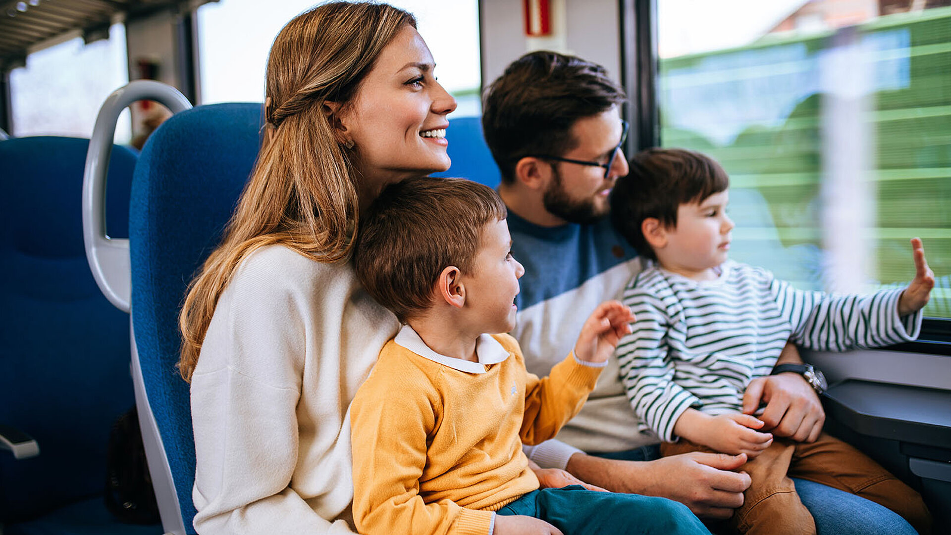 Familie sitzt im Zug und blickt gemeinsam aus dem Fenster, Kinder zeigen neugierig nach draußen.
