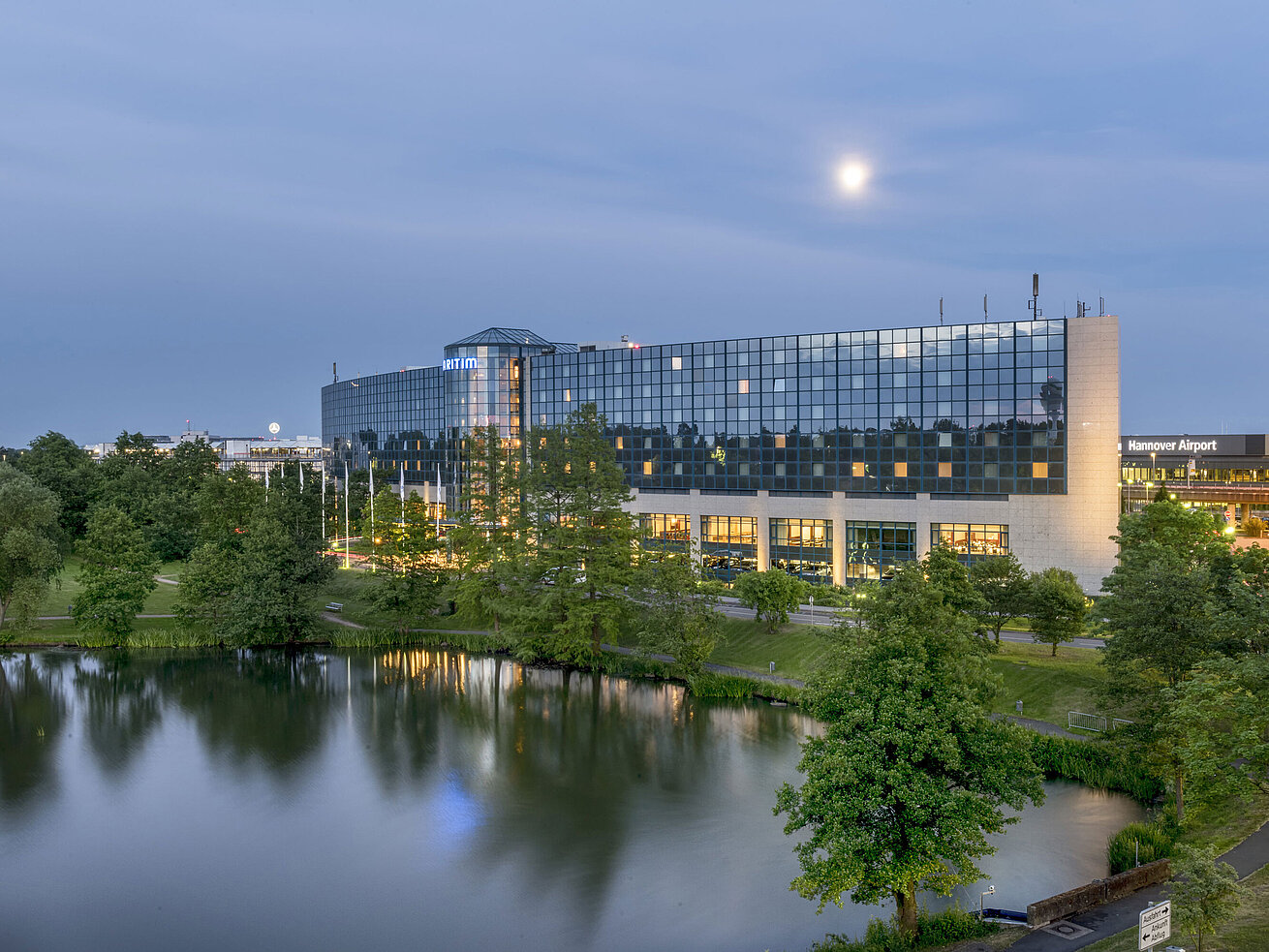 Beleuchtetes Maritim Hotel Hannover mit Glasfassade und Seespiegelung in der Abenddämmerung.