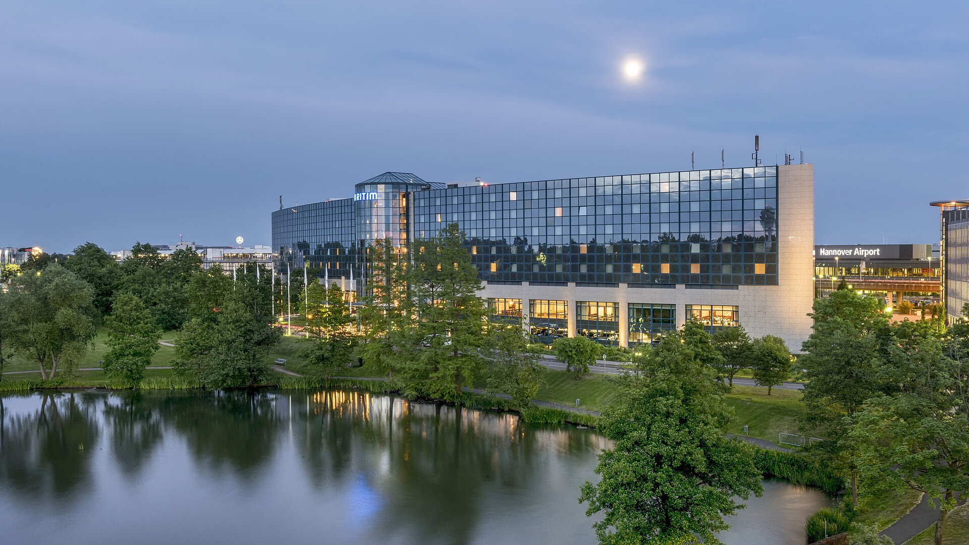 Beleuchtetes Maritim Hotel Hannover mit Glasfassade und Seespiegelung in der Abenddämmerung.