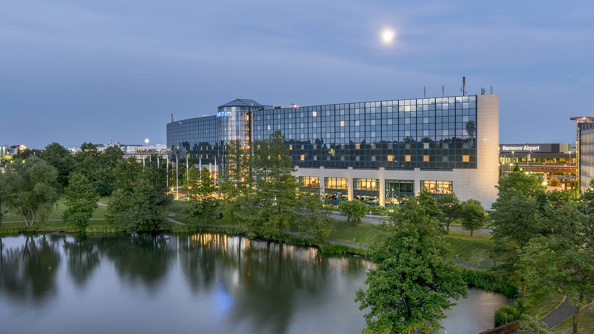 Beleuchtetes Maritim Hotel Hannover mit Glasfassade und Seespiegelung in der Abenddämmerung.