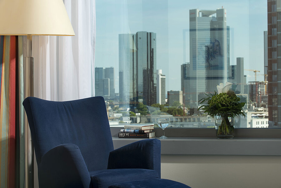 Ausblick auf die Skyline von Frankfurt Ausblick aus einer Suite im Maritim Hotel Frankfurt mit Skyline-Blick, blauem Sessel und dekorativem Blumenarrangement.