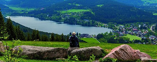 Wanderer mit Rucksack sitzt auf Felsen und schaut auf den Titisee und das Maritim Hotel im Schwarzwald