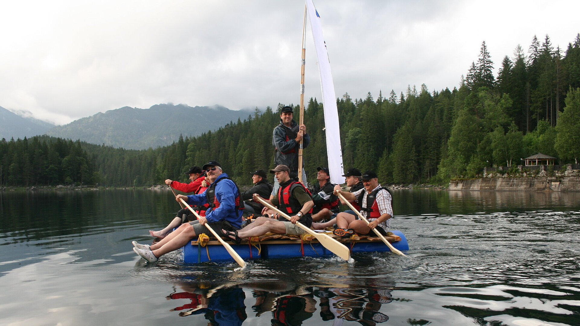 Gruppe paddelt auf Floß über ruhigen Bergsee vor Wald und Alpenlandschaft bei Outdoor-Teambuilding