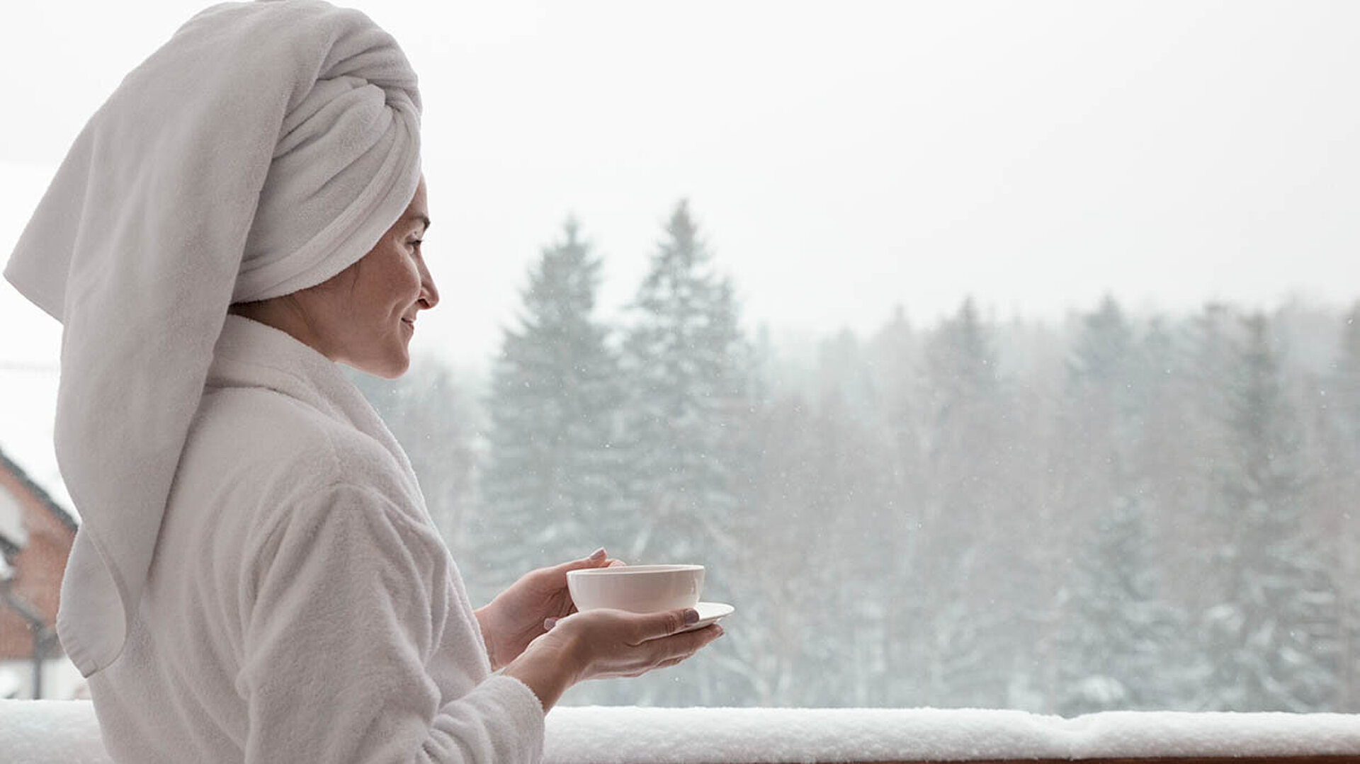 Person im Bademantel mit Handtuchturban hält eine Tasse und genießt den Blick auf verschneiten Wald.