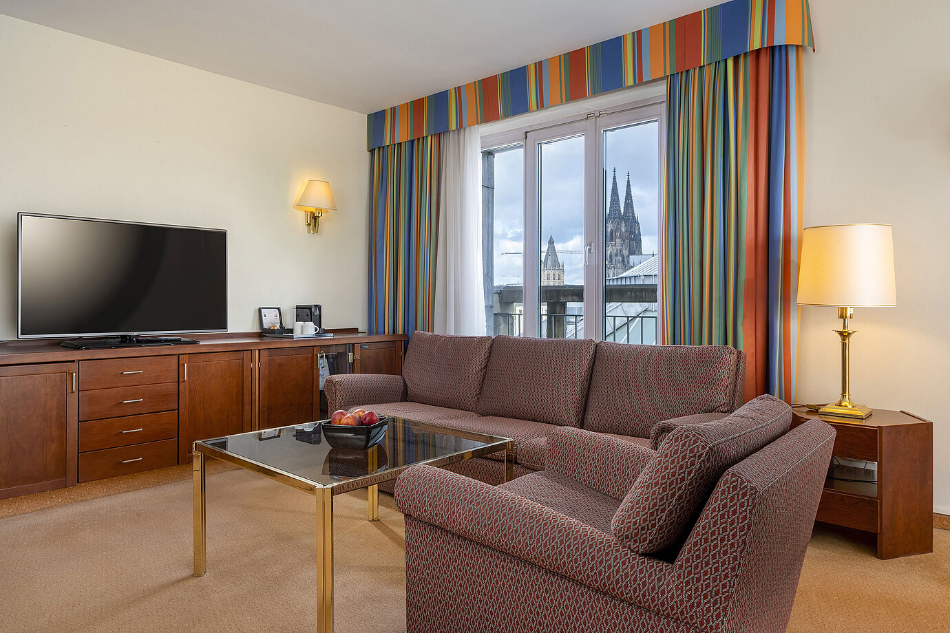 Wohnzimmer der Senior Suite im Maritim Hotel Köln mit Sofa, TV und Blick auf den Kölner Dom durch die Fenster.