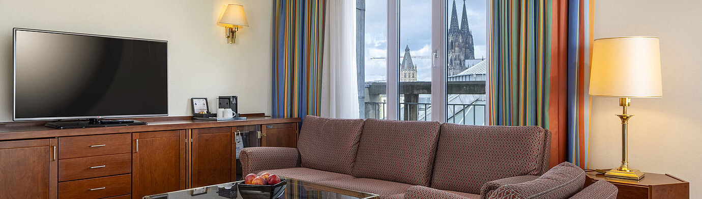 Wohnzimmer der Senior Suite im Maritim Hotel Köln mit Sofa, TV und Blick auf den Kölner Dom durch die Fenster.