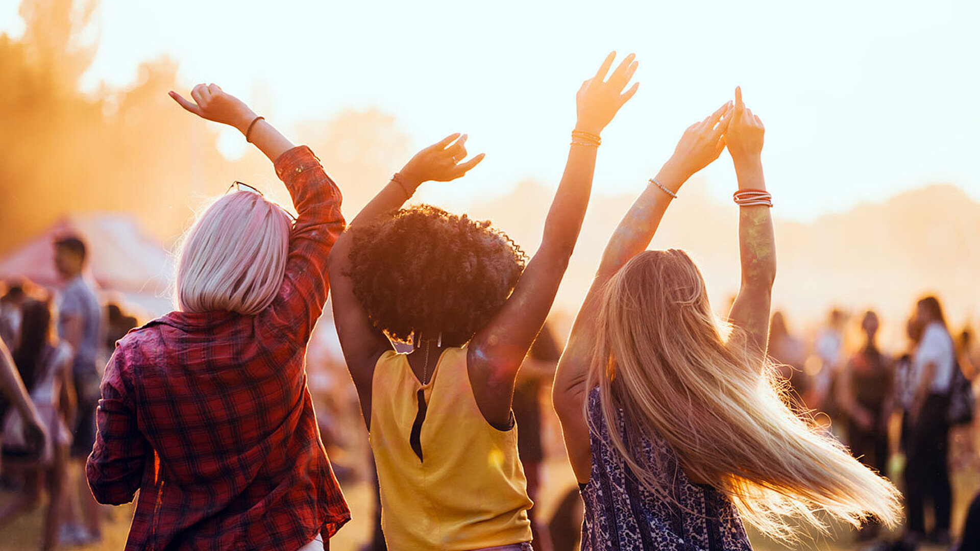 Drei Freunde tanzen mit erhobenen Armen im Sonnenuntergang bei einem Musikfestival im Freien.