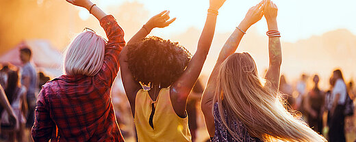 Drei Freunde tanzen mit erhobenen Armen im Sonnenuntergang bei einem Musikfestival im Freien.