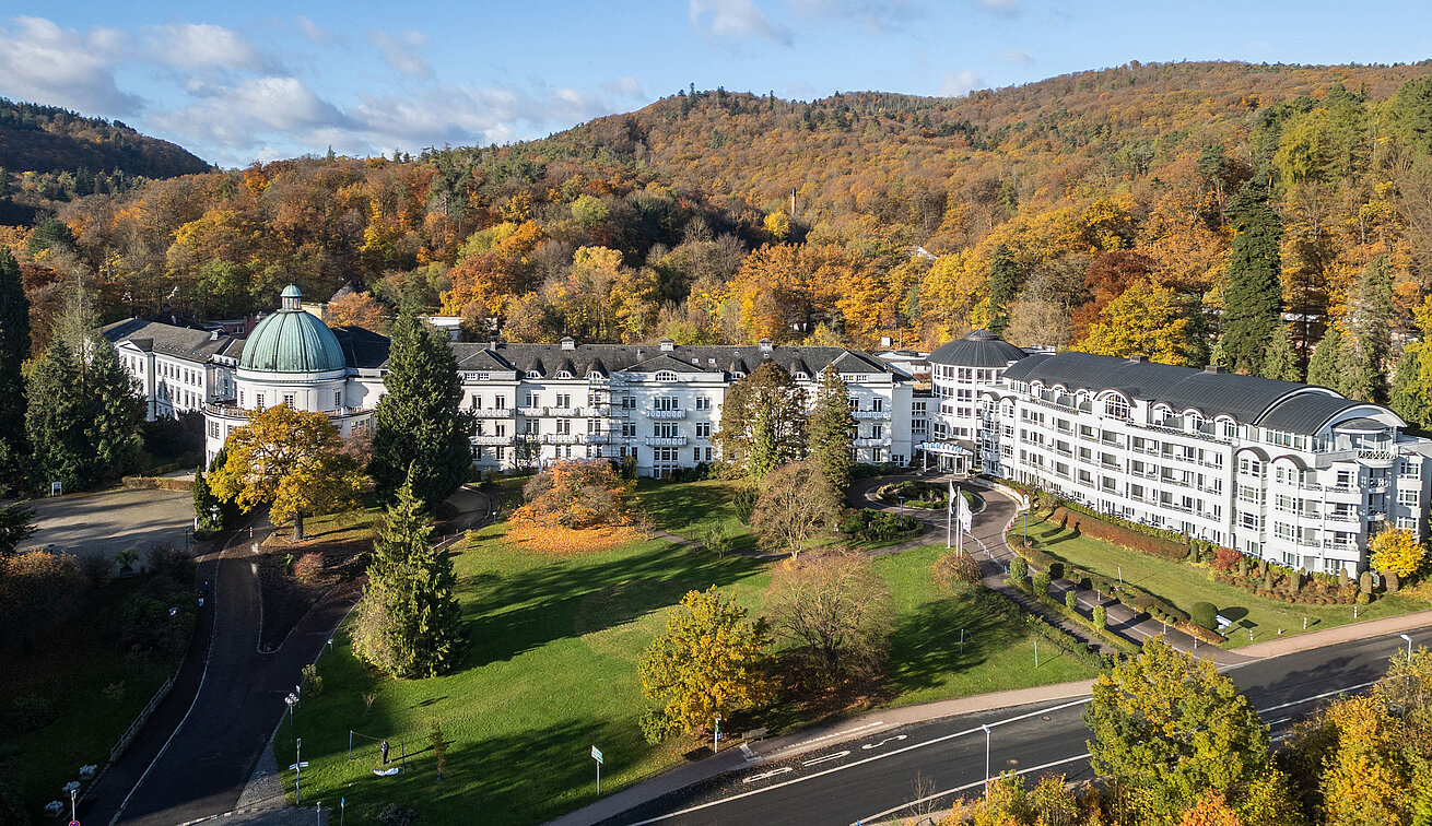 Außenansicht des Maritim Hotel Bad Wildungen mit Park, Bäumen und herbstlichem Wald im Hintergrund.