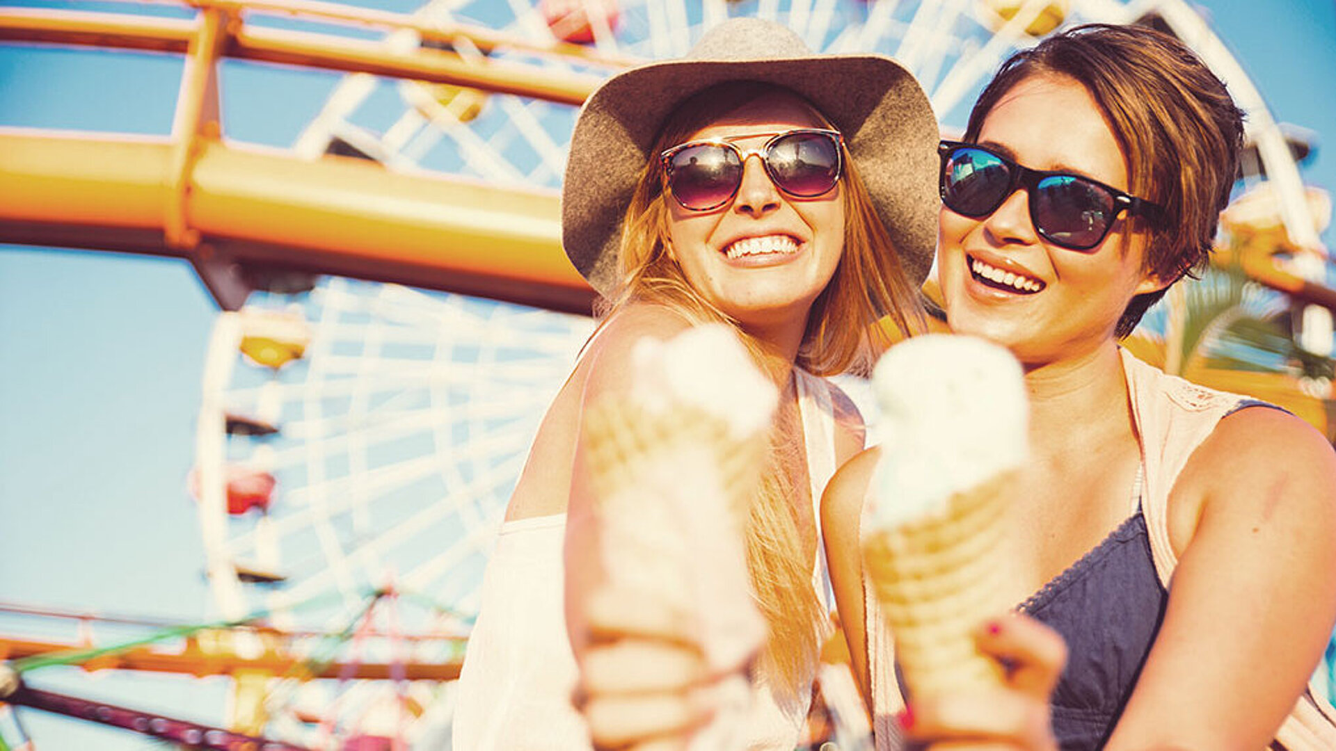 © oneinchpunch - AdobeStock.com Zwei Frauen essen lachend ein Eis. Im Hintergrund ist ein Riesenrad auf einem Jahrmarkt bei Sonnenschein zu sehen