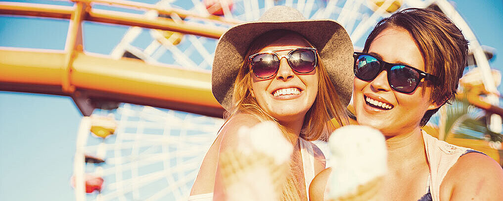 Zwei Frauen essen lachend ein Eis. Im Hintergrund ist ein Riesenrad auf einem Jahrmarkt bei Sonnenschein zu sehen