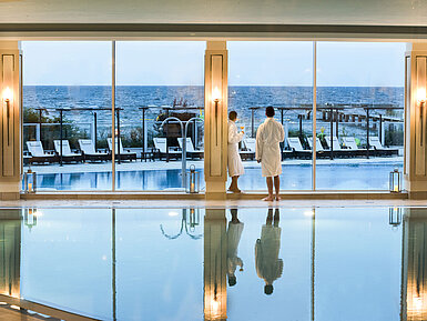 Schwimmbad  Innenpool mit Meerblick im Maritim Seehotel Timmendorfer Strand, zwei Gäste in Bademänteln am Fenster.