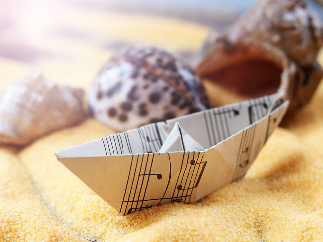 Gefaltetes Papierschiffchen auf dekorativem Sand mit Muscheln und maritimer Tischdekoration.